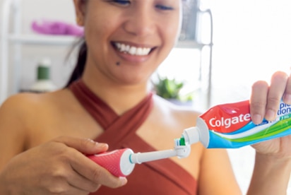 Woman smiling while adding Colgate toothpaste to a toothbrush in a bathroom.