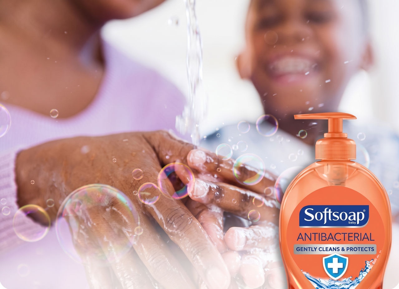 Close-up of hands washing with bubbles next to a Softsoap Antibacterial liquid hand soap bottle, with a smiling child blurred in the background.