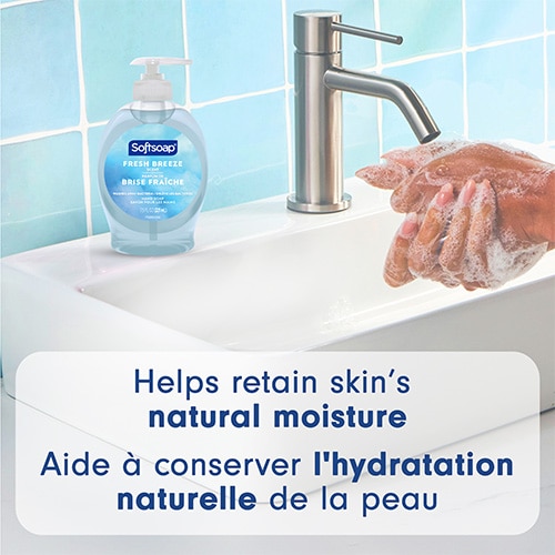 A Softsoap Fresh Breeze bottle sitting on a clean white vanity next to a faucet, with hands lathering soap. Text: "Helps retain skin’s natural moisture".