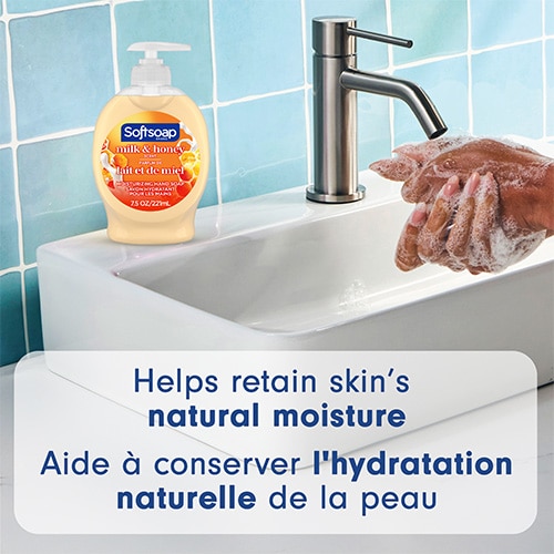 Softsoap Milk and Honey bottle sitting on a white bathroom sink next to a faucet, with hands lathering. Text: "Helps retain skin’s natural moisture".