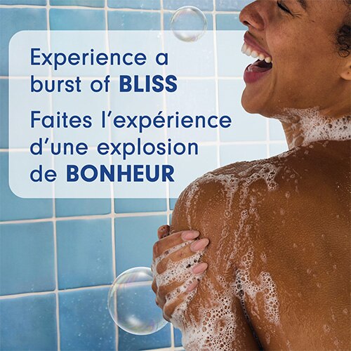 Un gros plan de style « lifestyle » montrant une personne souriant sous la douche, de la mousse de savon sur l'épaule. Un texte en surimpression indique : « Faites l’expérience d’une explosion de BONHEUR. »