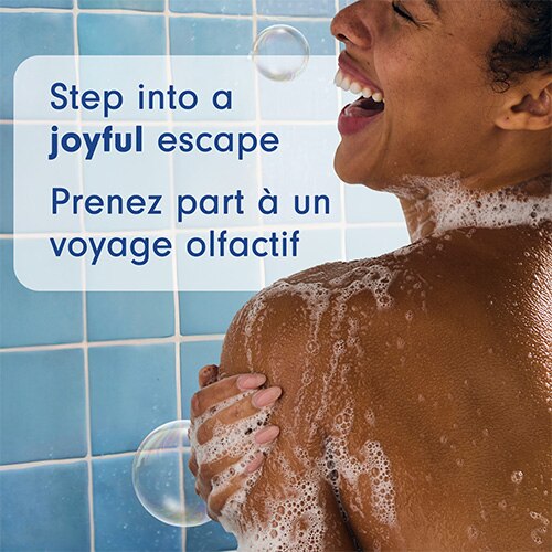 Une personne sourit sous la douche, de la mousse de savon sur l'épaule. Le texte en anglais et en français dit : Step into a joyful escape. Évadez-vous dans la joie.