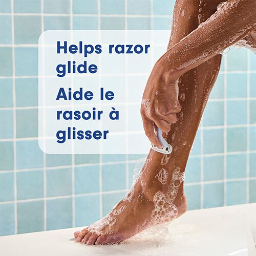 Gros plan sur une personne se rasant la jambe sous la douche avec une mousse rose onctueuse. Texte en anglais et en français : Helps razor glide, Aide le rasoir à glisser.