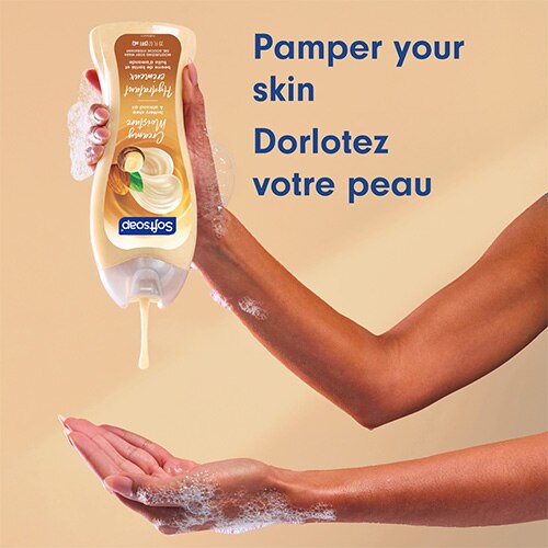 Une personne verse le gel douche crémeux, de couleur beige, de la bouteille dans sa paume. Le texte, en anglais et en français, indique : « Pamper your skin, Dorlotez votre peau ».