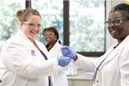 Deux scientifiques en sarrau et gants se tapent le poing en souriant; collègue flouté derrière au labo