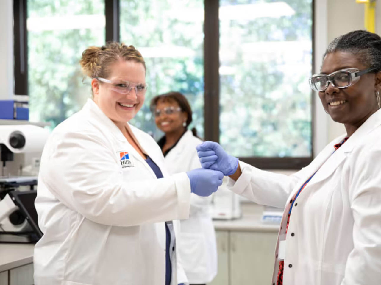 Deux scientifiques en sarrau et gants se tapent le poing en souriant; collègue flouté derrière au labo